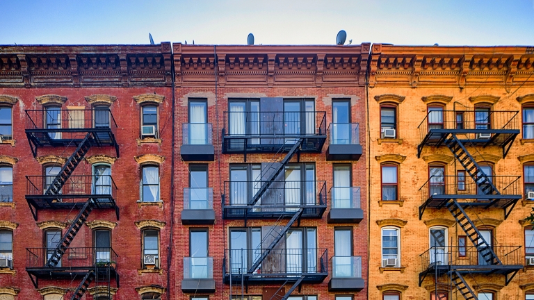 A row of vintage brick apartment buildings with fire escapes in front. The left building is red, the central one is a darker shade with modern balconies, and the right building is light orange. The sky is clear and blue.