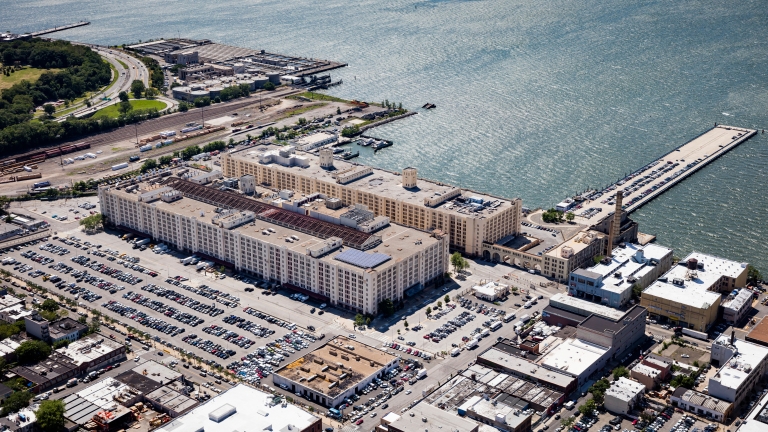 Aerial view of Brooklyn Army Terminal, a large waterfront warehouse complex with numerous parking lots filled with cars. The area is surrounded by industrial buildings, green spaces, and docks extending into the water.