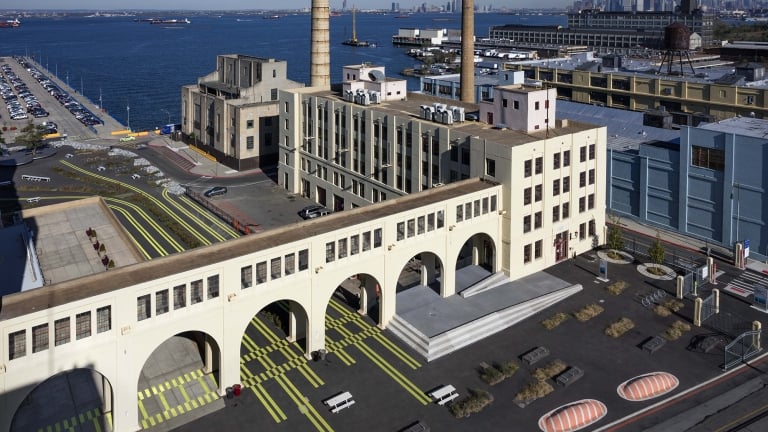 Aerial view of Brooklyn Army Terminal, an industrial waterfront complex with large, multi-story buildings and smokestacks. The area features pedestrian walkways with colorful lines, benches, and basketball courts. The skyline and water body are visible in the background. Aerial view of Brooklyn Army Terminal, an industrial waterfront complex with large, multi-story buildings and smokestacks. The area features pedestrian walkways with colorful lines, benches, and basketball courts. The skyline and water body are visible in the background.