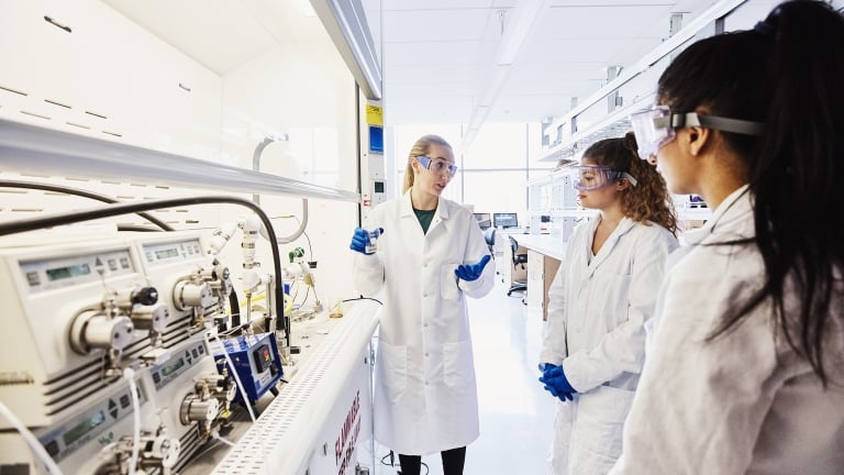 Three scientists wearing lab coats and safety goggles stand in a laboratory. One is holding a clipboard and gesturing while speaking to the others, who listen attentively. Various scientific equipment is visible on the counter beside them. Three scientists wearing lab coats and safety goggles stand in a laboratory. One is holding a clipboard and gesturing while speaking to the others, who listen attentively. Various scientific equipment is visible on the counter beside them.