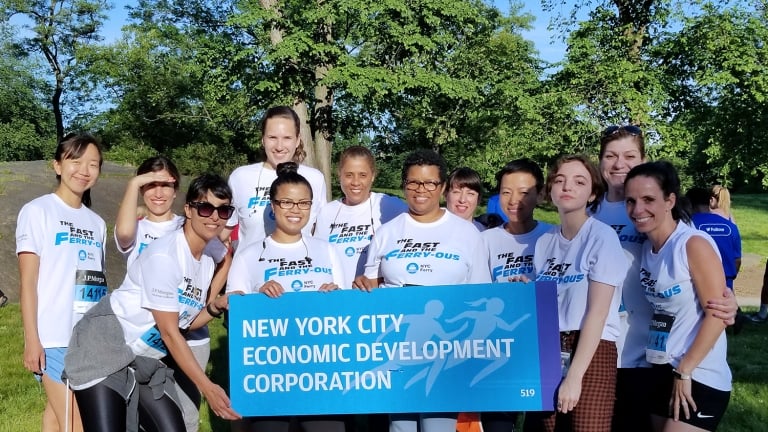 A group of people in matching t-shirts stand together outdoors on grass, smiling and holding a sign that reads New York City Economic Development Corporation. Trees and other participants are visible in the background.