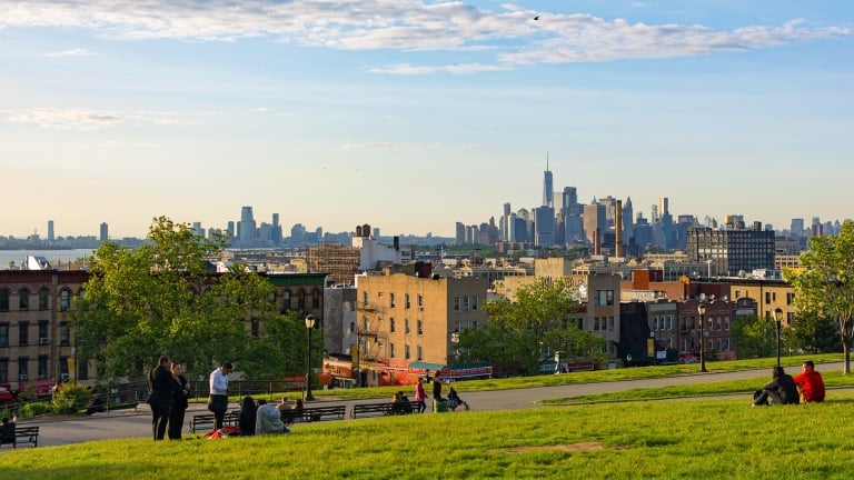 A grassy hill in Sunset Park, Brooklyn overlooking the New York City skyline at sunset. People sit on the grass and benches, enjoying the view. The sky is partly cloudy, with the sun casting warm light on the scene. Tall buildings are visible in the distance.