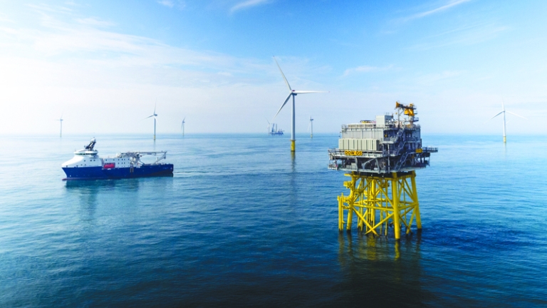 Offshore wind farm with a platform on yellow supports in the foreground and several wind turbines in the background. A vessel is visible on the left, surrounded by calm sea under a blue sky with scattered clouds.