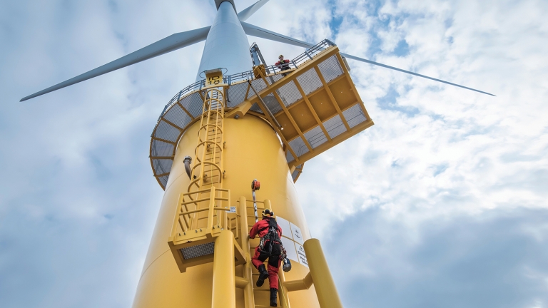 Show thumbnail preview A worker in red safety gear climbs a yellow ladder on a large offshore wind turbine tower. Another person is visible on a platform above. The sky is cloudy, and the turbine blades extend outward from the top.