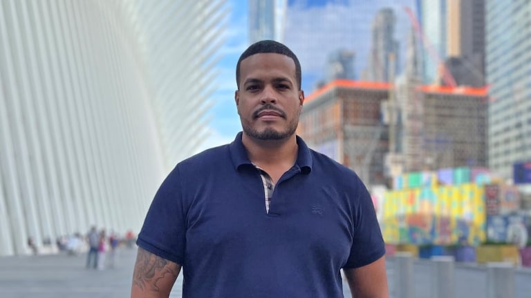 A man in a navy blue polo shirt, Joel Rivera, stands confidently with a modern architectural structure featuring white, rib-like beams behind him. The background includes skyscrapers and colorful construction site barriers. The sky is clear and blue. A man in a navy blue polo shirt, Joel Rivera, stands confidently with a modern architectural structure featuring white, rib-like beams behind him. The background includes skyscrapers and colorful construction site barriers. The sky is clear and blue.