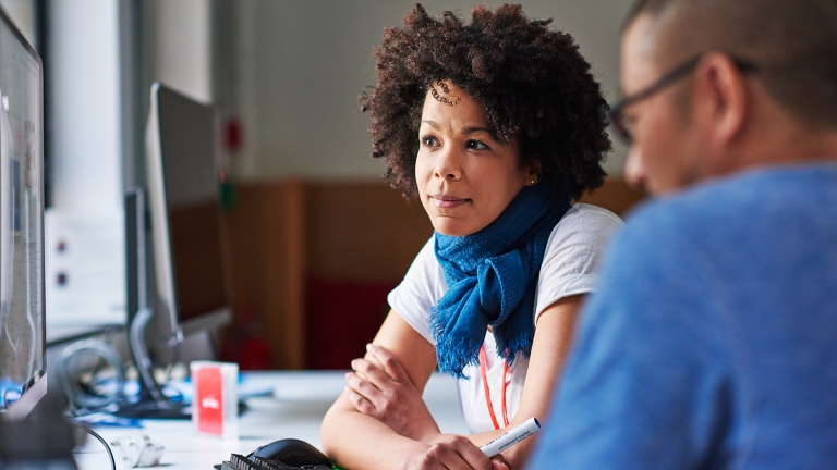 Image promoting Venture Access Alliance. A Black woman with curly hair and a blue scarf is attentively engaging with a man in a blue shirt. They are seated at a desk with computer monitors, suggesting a collaborative work environment.