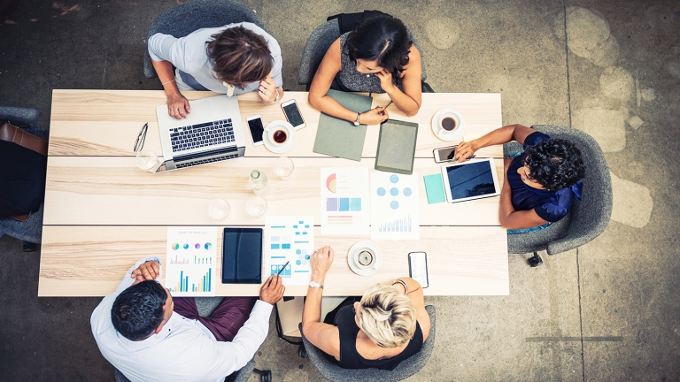 Image promoting Founder Fellowship program, part of Venture Access NYC. Five people are seated around a wooden table in an office setting. They are using laptops, tablets, and smartphones. There are also various charts and notebooks on the table, along with cups of coffee. The scene is viewed from above.