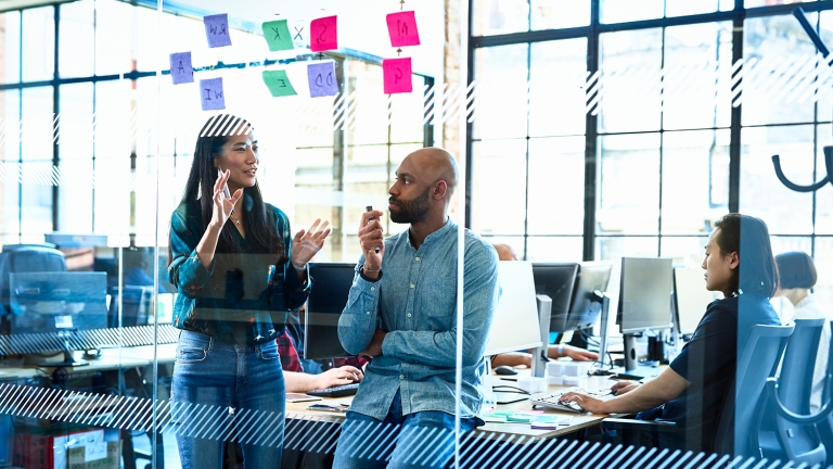 Header image of Venture Access NYC program page. A diverse group of people in a modern office setting. A woman is standing and gesturing while a man sits on a desk, listening. Another person works on a computer in the background. Colorful sticky notes are visible on the glass wall.