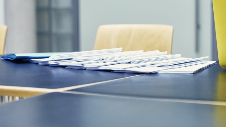 A stack of white envelopes is spread across a dark blue tabletop. Wooden chairs are partially visible in the background. The scene suggests a setting for organizing or distributing paperwork.