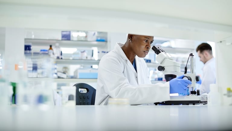 A scientist in a lab coat and blue gloves looks through a microscope in a laboratory. Shelves with equipment and another person working are visible in the background. The setting is bright and organized.