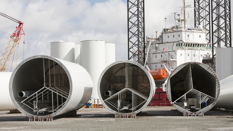 Three large cylindrical offshore wind turbine sections are laid horizontally on a construction site. Cranes and a ship are in the background under a partly cloudy sky. The scene suggests preparations for transportation or assembly.