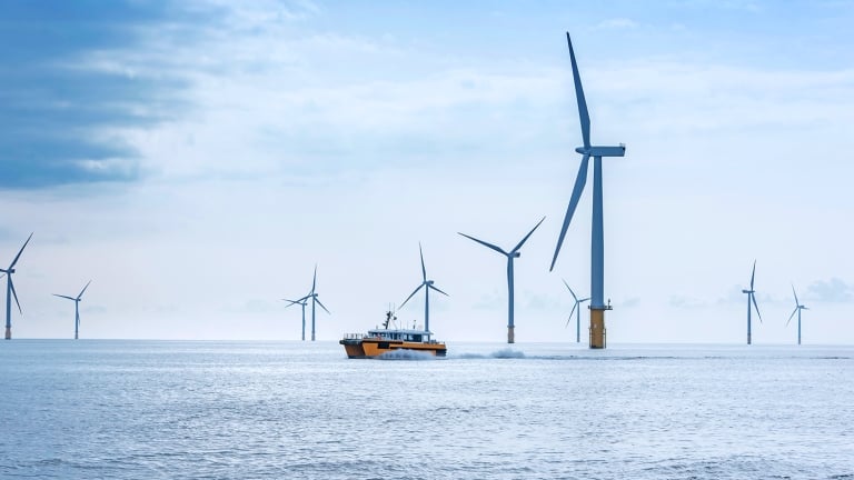 A boat navigates through a vast sea with numerous large wind turbines in the background, under a partly cloudy blue sky. The turbines are spaced out across the water, blending technological advancement with the natural seascape.