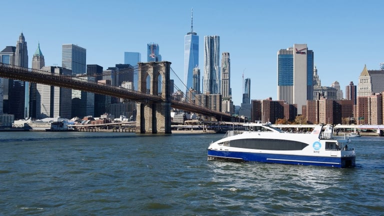 An NYC Ferry sails on the East River in front of the Brooklyn Bridge, with the New York City skyline towering in the background under a clear blue sky. Skyscrapers, including One World Trade Center, are visible.