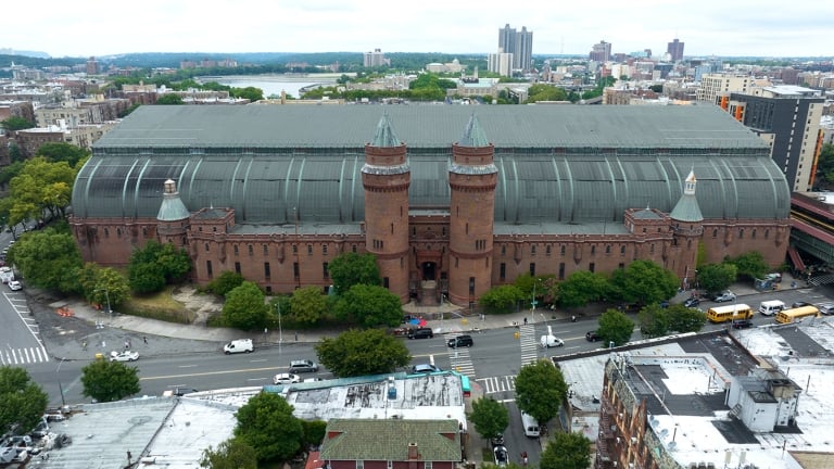 Aerial view of Kingsbridge Armory building with red brick walls and twin turrets, surrounded by NYC streets and greenery. The large, curved metal roof contrasts with the urban landscape, and several vehicles are visible on the roads nearby. Aerial view of Kingsbridge Armory building with red brick walls and twin turrets, surrounded by NYC streets and greenery. The large, curved metal roof contrasts with the urban landscape, and several vehicles are visible on the roads nearby.