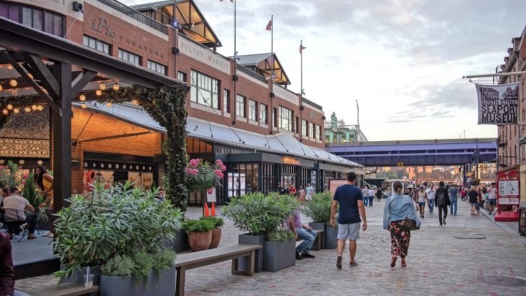 People walk along a cobblestone street by a historic market building with a Fulton Market sign. Outdoor seating with string lights and potted plants is on the left. Banners read South Street Seaport Museum. American flags are on the rooftops. People walk along a cobblestone street by a historic market building with a Fulton Market sign. Outdoor seating with string lights and potted plants is on the left. Banners read South Street Seaport Museum. American flags are on the rooftops.