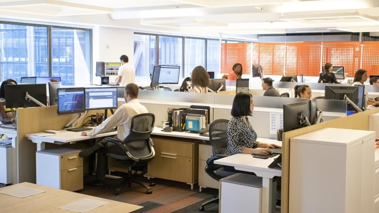 Open office space with people working at desks equipped with computers and monitors. Large windows and bright lighting create a spacious atmosphere. Some individuals are standing, while others are seated, all focused on their tasks.