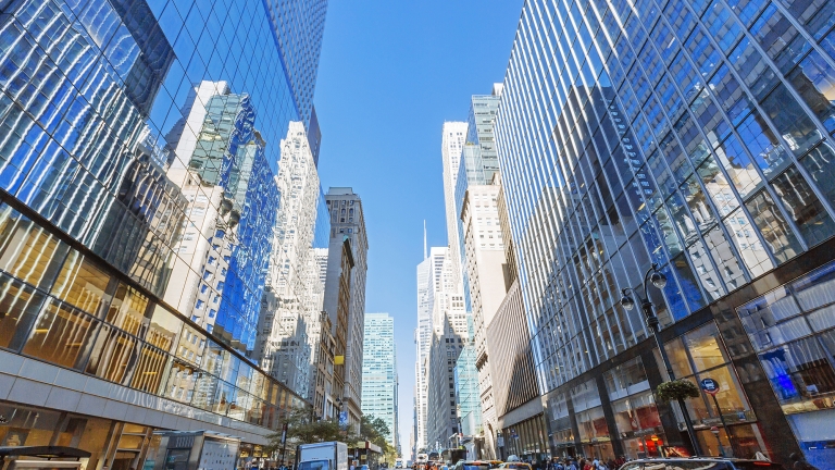 A vibrant city street flanked by tall skyscrapers with mirrored glass. The road is busy with vehicles, including cars and trucks, and pedestrians on the sidewalks. The sky is clear and blue, reflecting off the buildings.