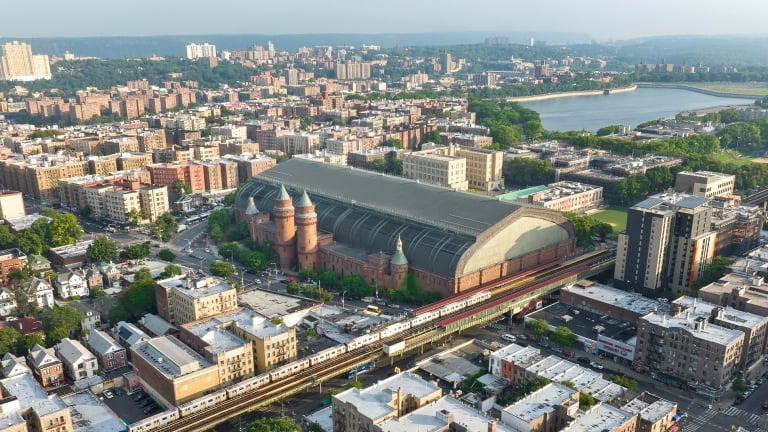 Rendering of Kingsbridge Armory. Aerial view of a cityscape featuring a large brick building with a curved glass roof, surrounded by residential and commercial buildings. A train is visible on tracks in the foreground, with greenery and a body of water in the background.