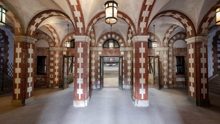 Interior of Kingsbridge Armory shows a grand hallway with vaulted ceilings and brick arches. Three hanging lanterns illuminate the space, and three open doorways lead to an outdoor area. The floor and walls feature a mix of brick and stone detailing.