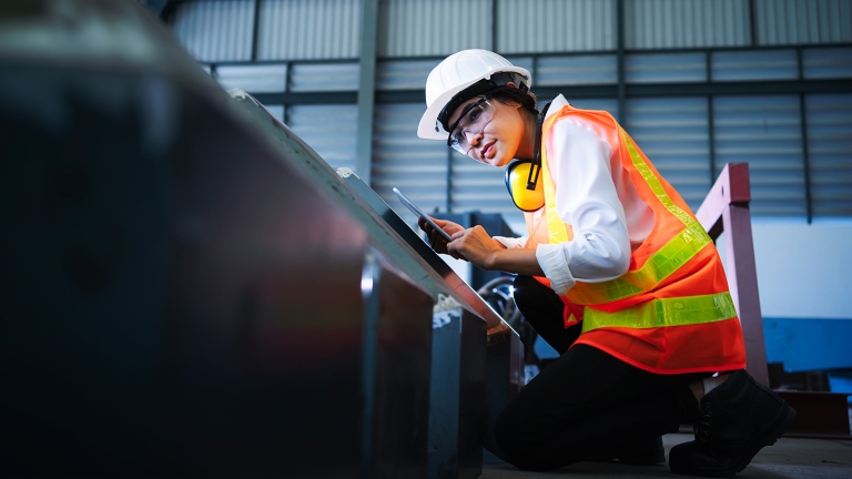 An Offshore Wind NYC Waterfront Pathways Program female engineer inspector wearing a white hard hat, orange safety vest, and yellow ear protection kneels beside industrial equipment indoors. She is holding a tablet and appears focused, with a corrugated metal wall in the background.