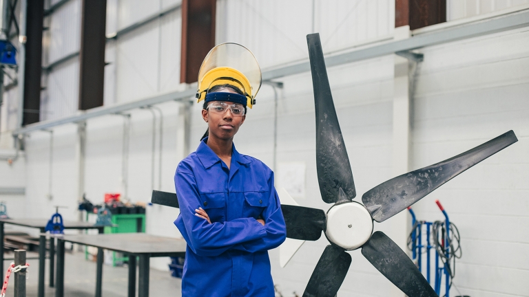 A woman in blue work attire and safety gear stands with arms crossed in an industrial setting, next to a large fan blade. The background shows workbenches and equipment, indicating a workshop or manufacturing environment.