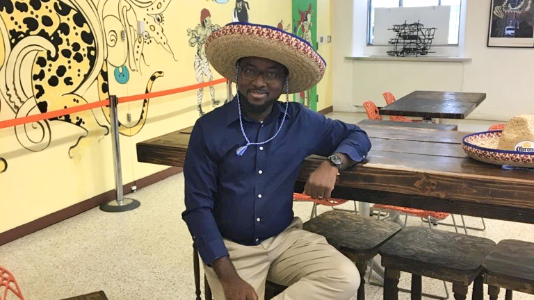 Sakiru Okeowo wearing a large straw sombrero sits casually at a wooden table in a vibrant room with colorful murals and a rope barrier. He is dressed in a blue shirt and beige pants, with a relaxed expression. Sakiru Okeowo wearing a large straw sombrero sits casually at a wooden table in a vibrant room with colorful murals and a rope barrier. He is dressed in a blue shirt and beige pants, with a relaxed expression.