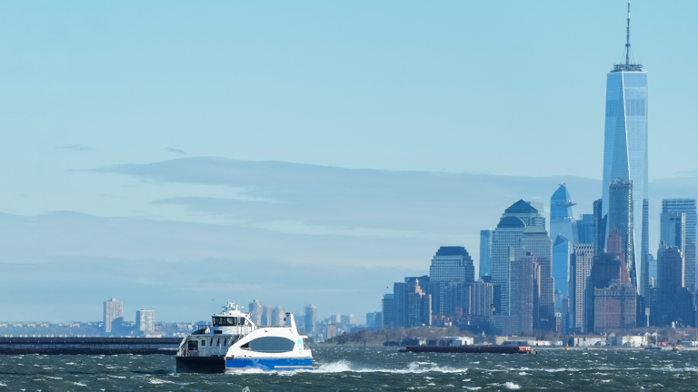 White and blue NYC Ferry travels across choppy water with the New York City skyline, including the One World Trade Center, visible in the background under a clear sky. White and blue NYC Ferry travels across choppy water with the New York City skyline, including the One World Trade Center, visible in the background under a clear sky.