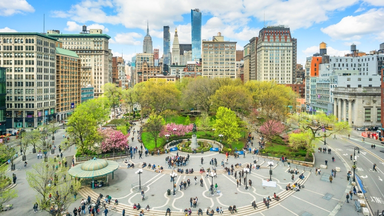 Aerial view of New York&#039;s Union Square, a lively urban park surrounded by tall buildings under a blue sky with clouds. The park features lush green trees, blooming pink flowers, a central fountain, and numerous people walking and sitting.