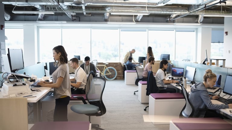 A modern real estate office with several people working at desks equipped with computers. Some are seated while others stand at adjustable desks. Large windows allow natural light in, and theres a bicycle near the windows. The decor is minimal and modern.