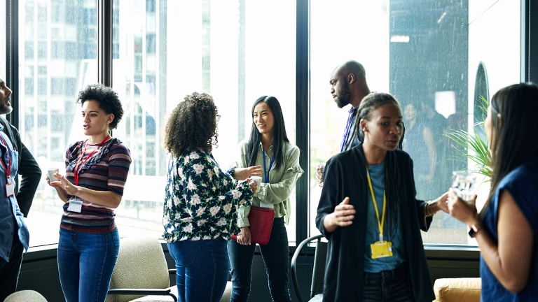 A group of people participating in NYCEDC Fellowship Program stand in a brightly lit room with large windows, engaging in conversation. They are casually dressed and appear to be in an office or conference setting. Coffee cups are on a table nearby.