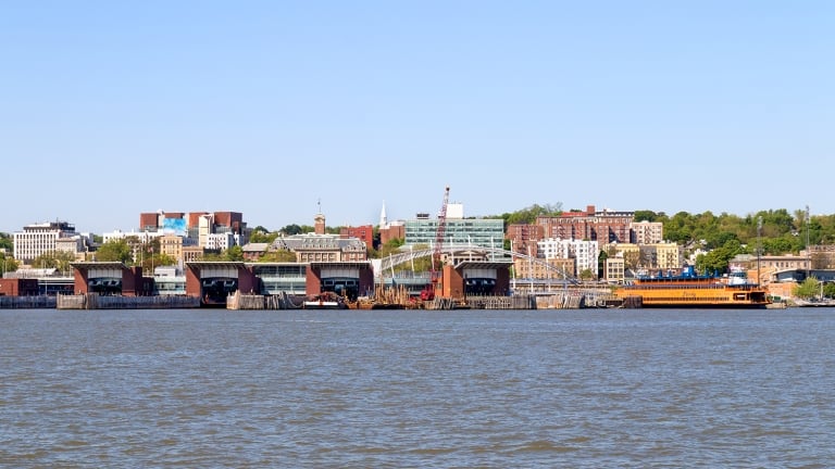 View of St. George Esplanade, Staten Island, New York City with several ferry terminals along the water. Buildings of various sizes and colors are scattered on a hill in the background under a clear blue sky.