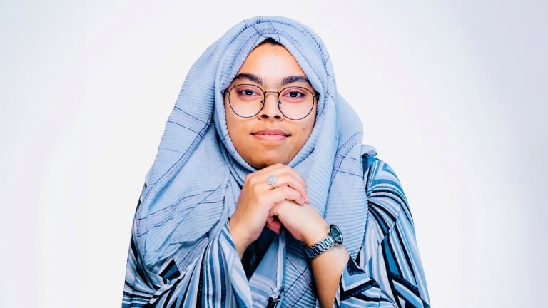 A female Cyber NYC intern, Samira Akram, wearing glasses and a light gray hijab, resting her chin on her hands. She is wearing a striped outfit and a watch, and is looking directly at the camera with a neutral expression against a plain background. A female Cyber NYC intern, Samira Akram, wearing glasses and a light gray hijab, resting her chin on her hands. She is wearing a striped outfit and a watch, and is looking directly at the camera with a neutral expression against a plain background.