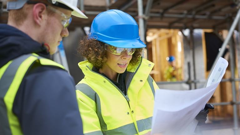 A female construction worker stands on a building site housing development and instructs a co-worker.