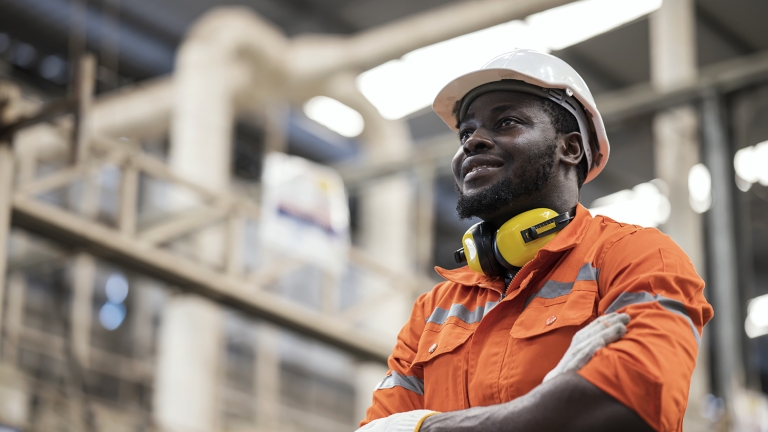 A man in an orange safety uniform and white hard hat stands with arms crossed in an industrial setting. He wears yellow ear protection around his neck and white gloves, looking upwards with a confident expression. Pipes and machinery are in the background.