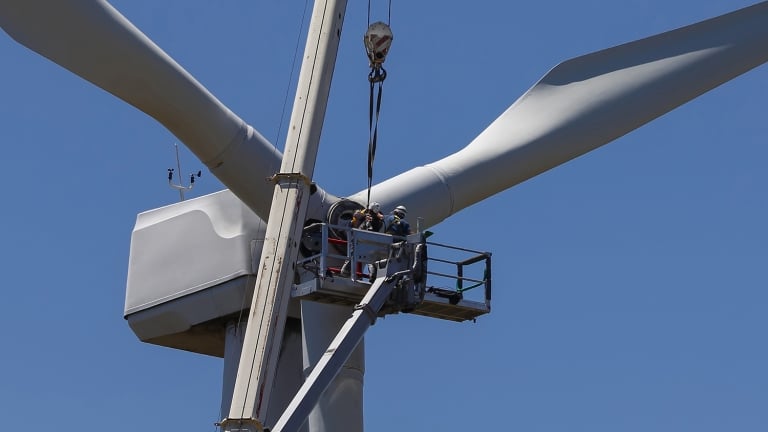 Workers in a crane basket perform maintenance on the blades of a large wind turbine against a clear blue sky. Workers in a crane basket perform maintenance on the blades of a large wind turbine against a clear blue sky.