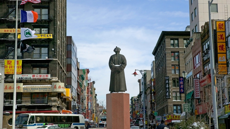 Ze Xu Statue stands in Chinatown, NYC, surrounded with buildings featuring Chinese signs. Multiple flags, including the U.S. flag, are visible, with pedestrians and vehicles in the background under a blue sky.
