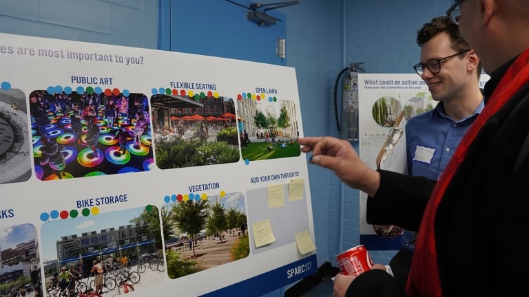 Two people view a display board with images and text about urban planning features like public art, seating, and vegetation. Sticky notes are visible for adding thoughts. They are having a discussion, one holding a drink can.