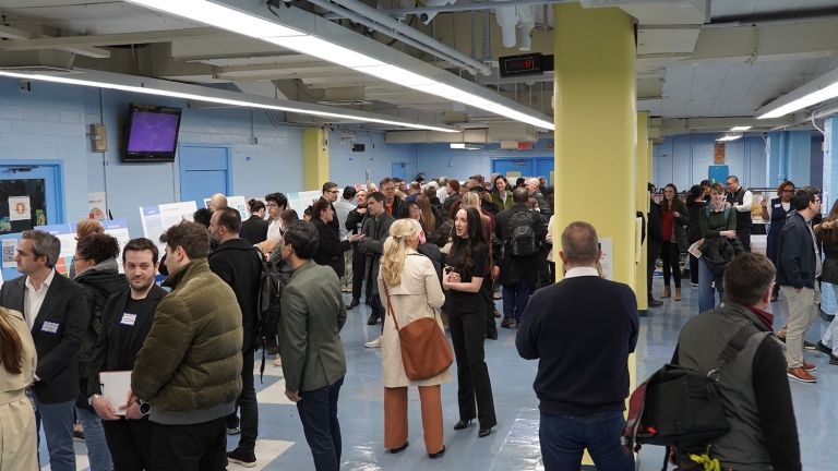 A large group of people gather in a room with blue walls and yellow pillars at a SPARC Community Engagement meeting. They appear to be engaged in conversations, some standing around tables with papers and displays. Overhead lights illuminate the space, creating a busy, social atmosphere.