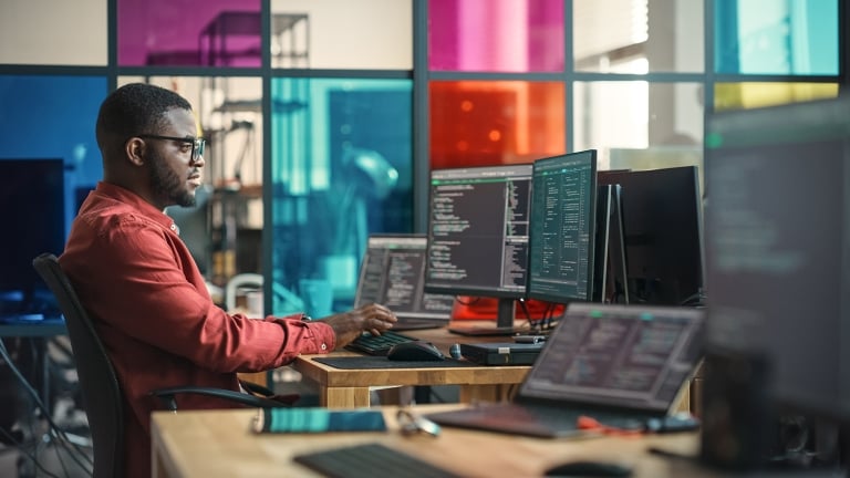 African American Man Writing Lines Of Code On Desktop Computer With Multiple Monitors and Laptop in Creative Office..