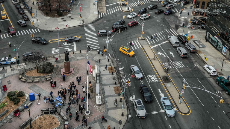 Aerial view of Chatham/Kimlau Square in New York City's Chinatown. A busy intersection with many cars, taxis, and people crossing the street. A small park with a statue and American flags is visible in the lower left corner. Multiple crosswalks and traffic signals are present.