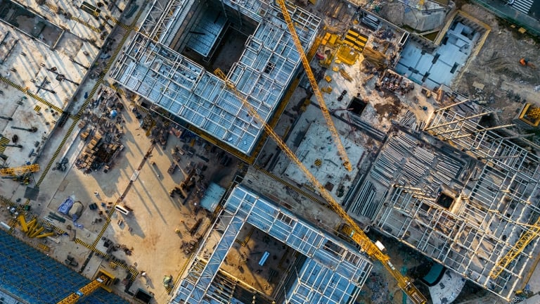 Aerial view of a construction site with multiple buildings in progress. Steel frameworks and construction cranes are visible, along with scattered materials and equipment. The site is busy with activity, under clear weather conditions.