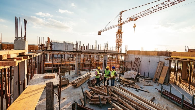 Three construction workers in safety gear discuss plans at a building site during sunset. Surrounding them are piles of wood, metal bars, and a crane, indicating ongoing construction. The skyline features a mix of clouds and blue sky.