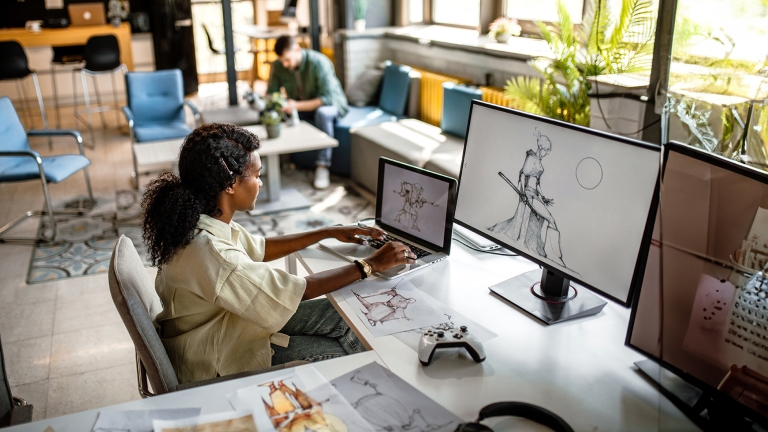 Young, black video game designer in company studio, working at her desk computer, drawing new sketches.