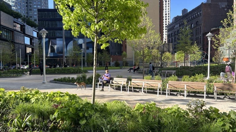 Abolitionist Place Park in Brooklyn with green trees, people sitting on benches, dogs on leash, and high-rise buildings in the background.