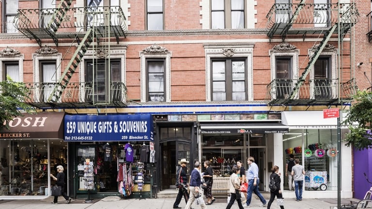 A group of people walk past storefronts, including a gift shop and a clothing store, on Bleecker Street with brick buildings and fire escapes.