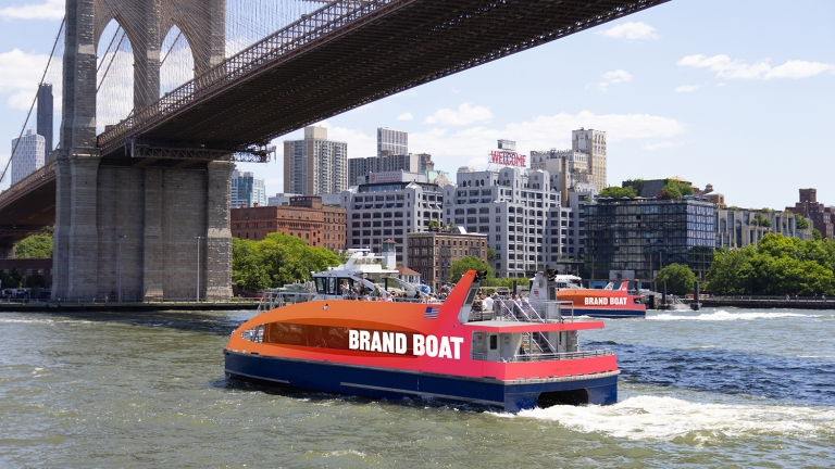 An orange and blue ferry labeled &quot;BRAND BOAT&quot; cruises under the Brooklyn Bridge with the New York City skyline in the background on a sunny day.