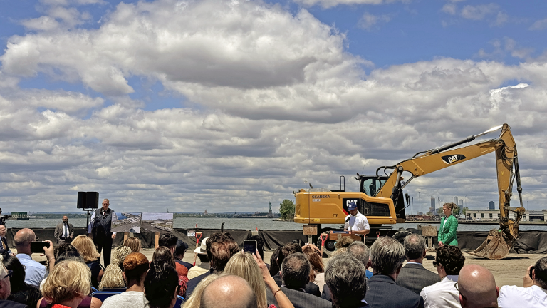 A group of people listening to NYC Mayor Eric Adams speaking at the South Brooklyn Marine Terminal groundbreaking ceremony with a large excavator in the background and a partly cloudy sky.