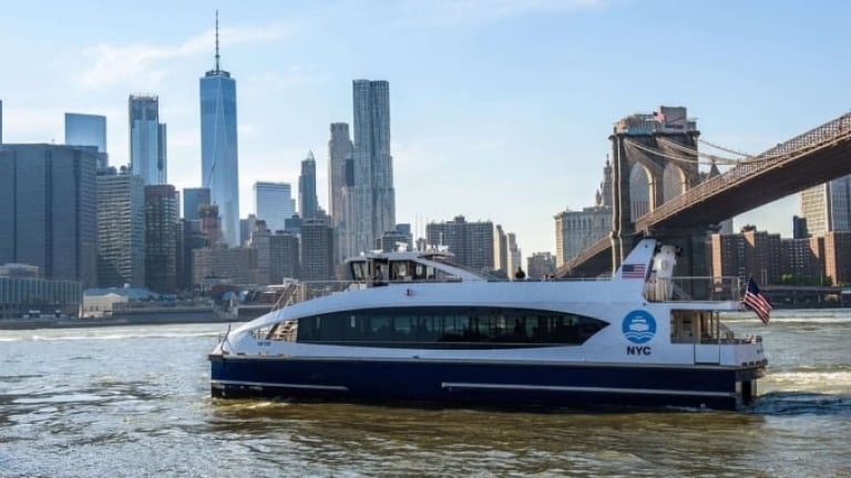 NYC Ferry sails on the river with the Manhattan skyline and Brooklyn Bridge in the background on a clear day.