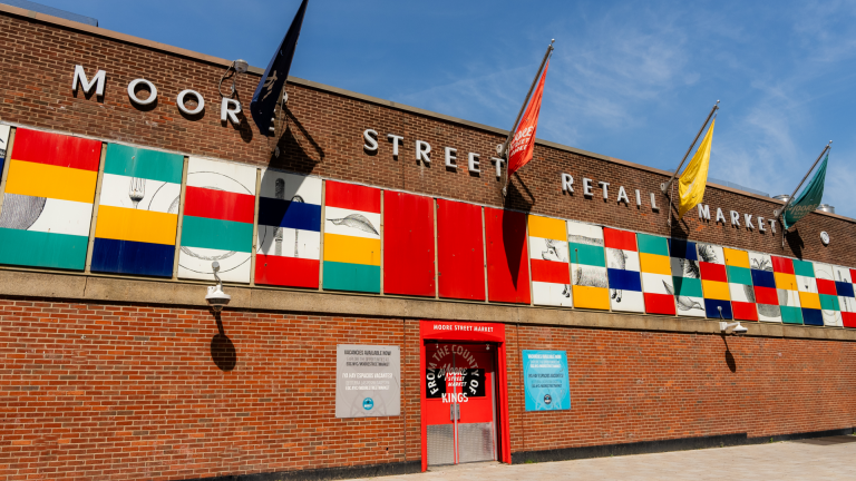 Exterior of Moore Street Retail Market, a brick building with colorful geometric panels and various flags. The entrance is marked by a red frame, flanked by informational signs. The sky is clear and blue.