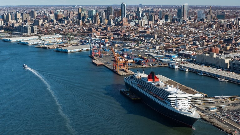 Aerial view of a docked cruise ship at the Brooklyn Cruise Terminal with the New York City skyline in the background.